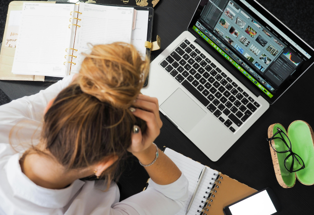 This overhead shot depicts a person with a bun hairstyle wearing a white shirt, working at a desk. The person is propping their head up with their hands while looking at a laptop displaying various designs. To the left of the computer, eyeglasses are placed in an eyeglass case, and to the right, an open coil-bound book displays notes.
