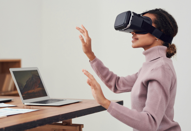 A woman in a pink sweater sits at a desk using a VR headset. Her hands are raised and waving in the air, indicating she is engaged with the virtual reality experience. An open laptop sits on the desk in front of her.