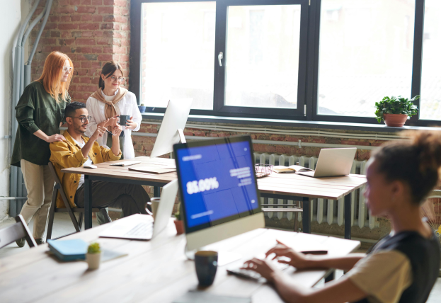 This scene depicts an office with two desks facing each other, sunlight streaming through the windows. In the foreground, a person with a bun hairstyle is typing on a desktop computer. Across the room, three colleagues are gathered around another desktop computer. Two are standing, one holding a coffee mug, and the third person is sitting, all engaged in discussion.