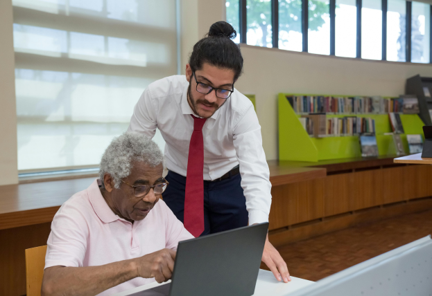 Image Description: The image depicts an older man seated at a table, actively using a laptop, while a younger man in a white shirt and red tie leans over, providing guidance. The setting is a modern library, featuring bright lighting, large windows, and shelves filled with books.