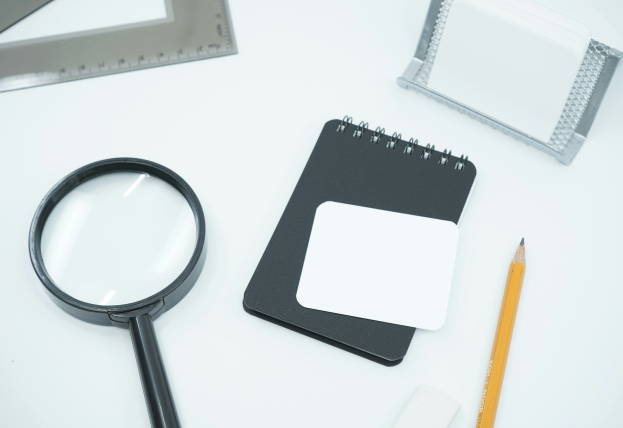 Image Description: The image features a flat lay arrangement on a white surface, including a magnifying glass, a black spiral-bound notepad with a blank white card on top, a pencil, a white eraser, a metallic cardholder, and a triangular ruler.