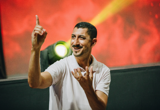 The image features a man in a white polo shirt smiling and signing. He appears to be on a stage with a dramatic red and yellow backdrop and a spotlight behind him.
