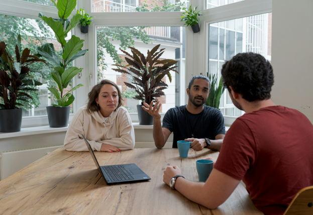 Three people are sitting around a wooden table in a well-lit, modern workspace with large windows and greenery in the background. A laptop is open on the table, and two individuals are holding blue coffee cups. One person, wearing a black shirt, is actively speaking and gesturing with his hand, while the other two, dressed in a beige hoodie and a brown shirt, are attentively listening.