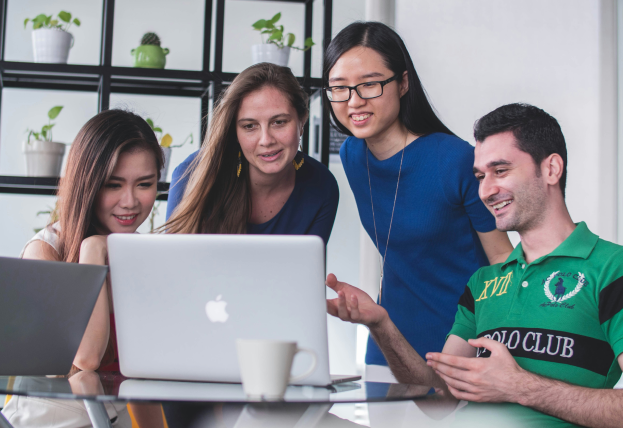 The image shows four diverse young professionals gathered around a laptop, engaged in a discussion. They appear to be collaborating on a project, with one person gesturing while speaking and the others attentively looking at the screen. The setting is a modern office with shelves decorated with small potted plants.