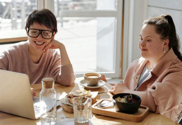 Two women sitting at a café table, smiling and enjoying a meal while looking at a laptop.